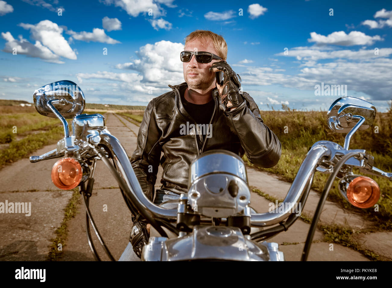 Biker parlando su uno smartphone. Biker uomo che indossa una giacca di pelle e occhiali da sole seduto sulla sua moto. Foto Stock