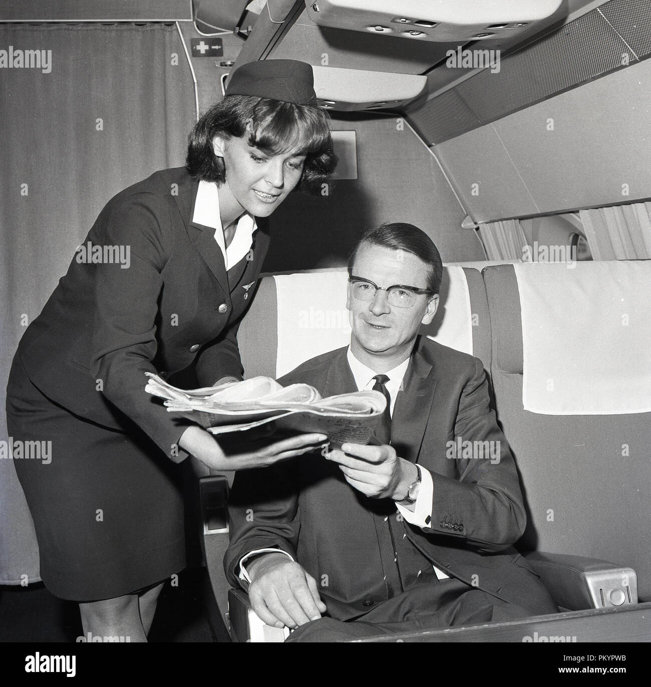 Anni sessanta, storico, una femmina in uniforme assistente di volo presentando un quotidiano per un maschio adatto passeggero seduto in poltrona Business Class a bordo di un aereo. Foto Stock