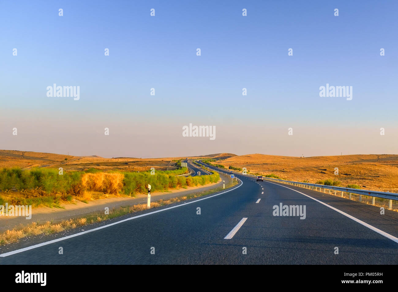 La guida lungo vuoto strade tortuose in Andalusia, Spagna Estremadura vicino alla fine della stagione estiva con un golden sunrise illuminazione i campi a secco ed un bl Foto Stock