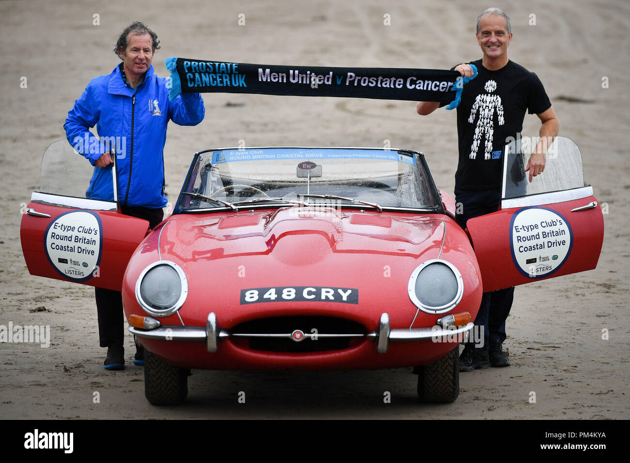 Nipote di Sir Malcolm Campbell MBE, Don Galles (destra) e Jaguar conducente Philip porter mantenere una sciarpa a sostegno del cancro della prostata UK carità oltre l'E-type Jaguar che è stata usata nel film Italian Job come dozzine di E-tipi iniziano a coastal intorno al Regno Unito viaggio in aiuto del cancro della prostata la carità a partire da Pendine Sands, Carmarthenshire, Galles. Foto Stock