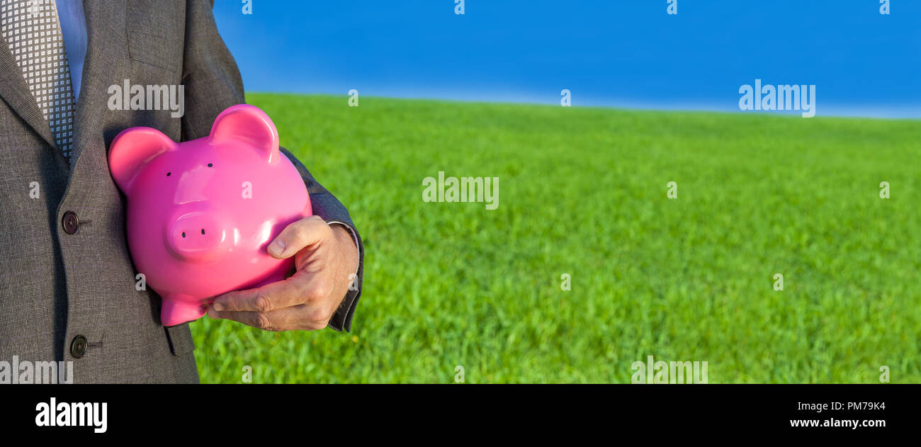 Panoramica banner web verde il concetto di investimento colpo di un uomo o di imprenditore nel soddisfare tenendo un rosa salvadanaio in un campo verde con un luminoso cielo blu. Foto Stock