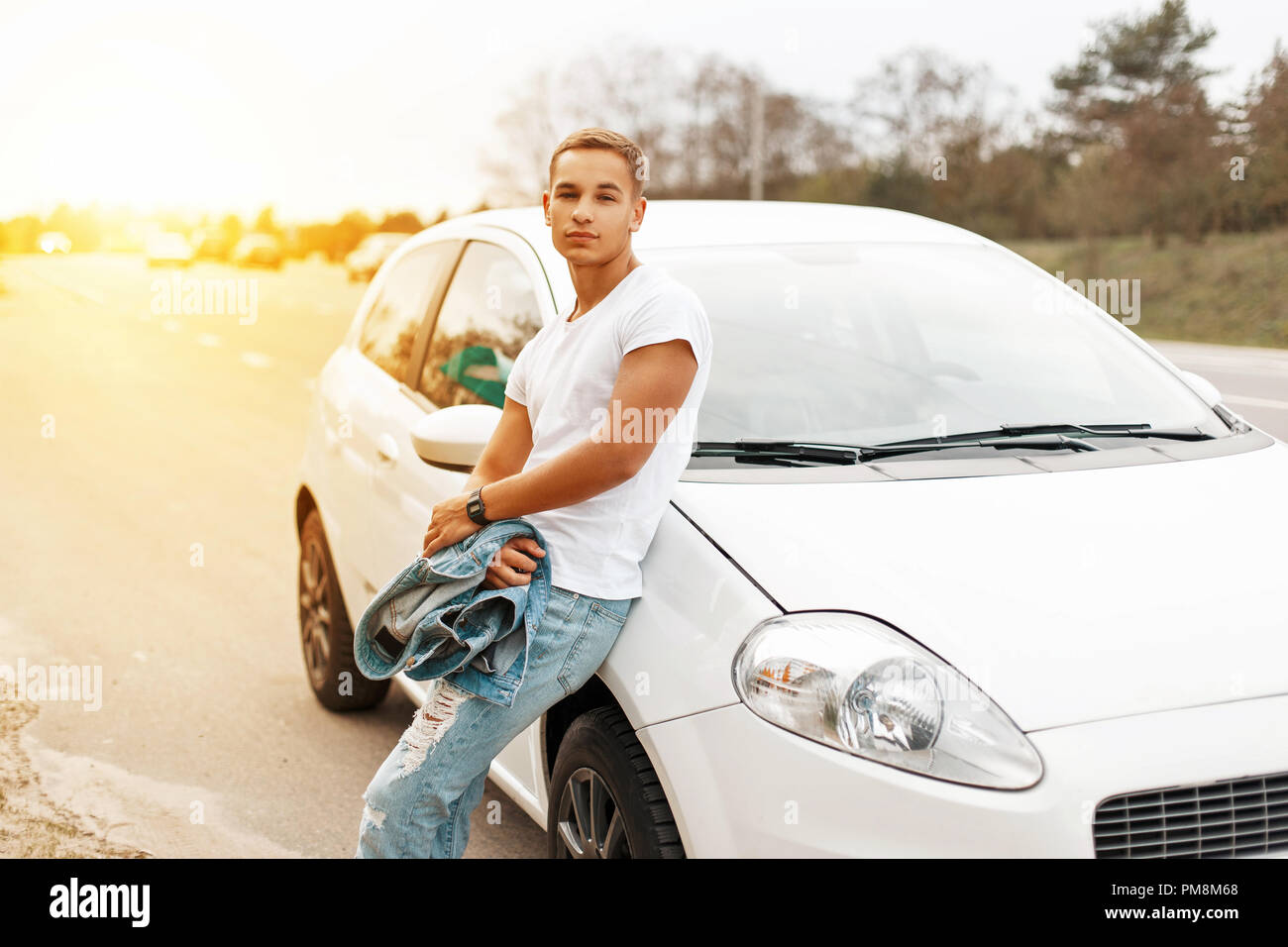 Bel giovane in un bianco tank top vicino la vettura al tramonto. Viaggiare sulle strade Foto Stock