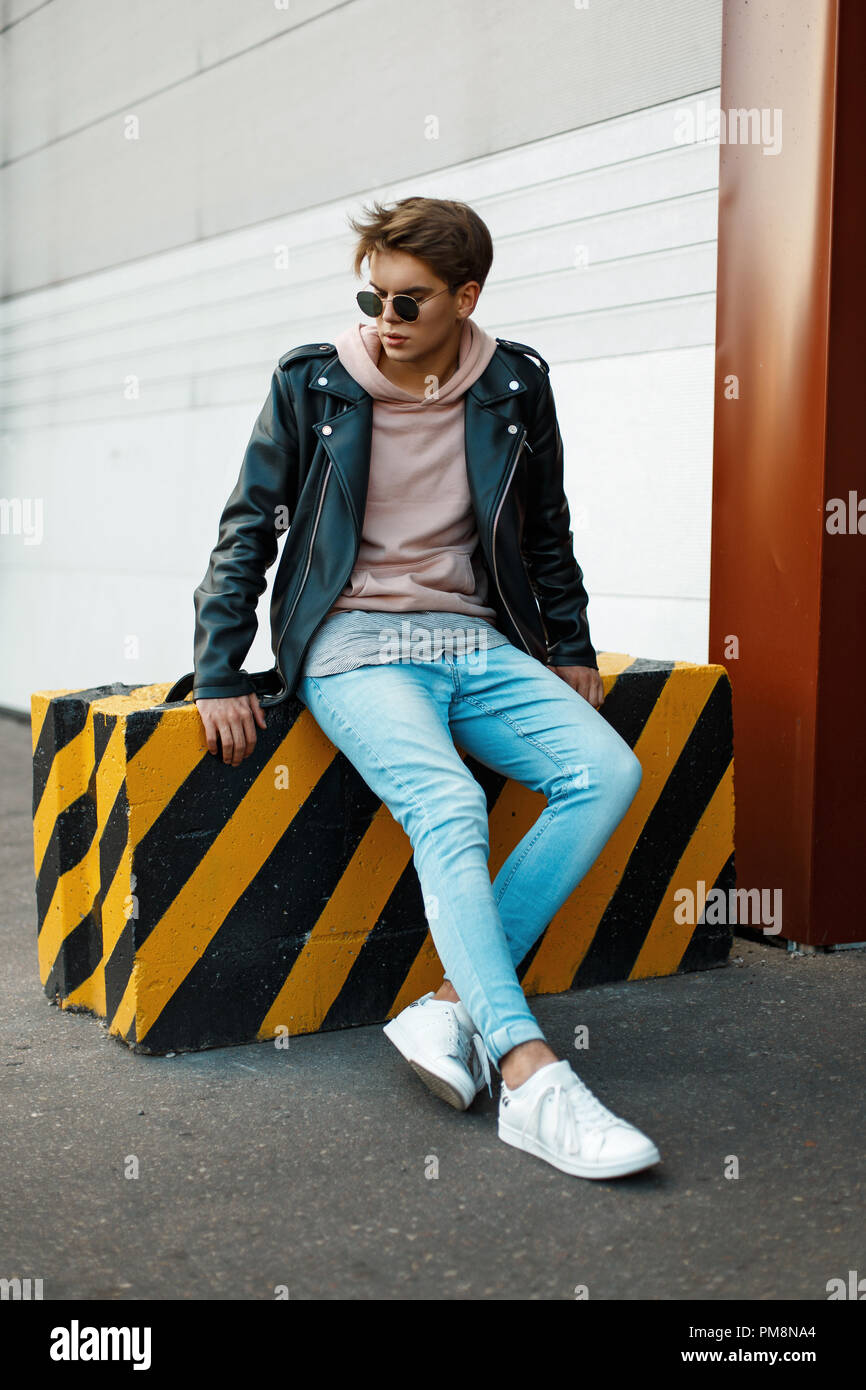 Bel giovane con occhiali da sole in un nero giacca di pelle, jeans blu e  bianco sneakers siede su un giallo-nera lastra Foto stock - Alamy