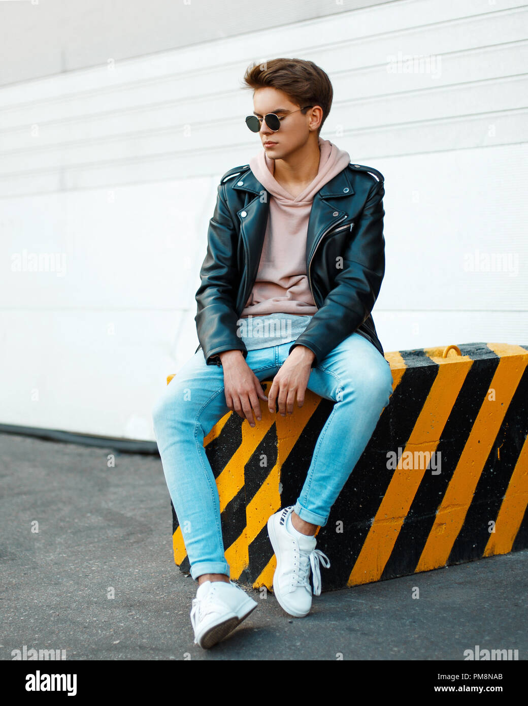 Giovane uomo bello con occhiali da sole in un nero elegante giacca di  pelle, una felpa rosa, blu jeans e scarpe bianche si siede su un giallo-nera  lastra Foto stock - Alamy