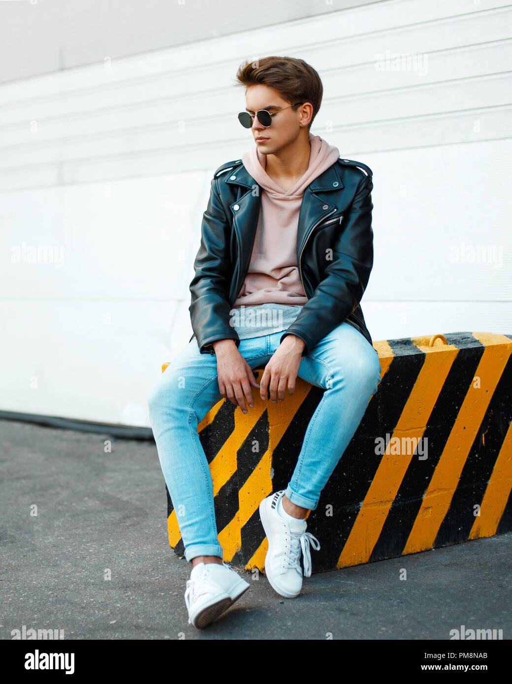 Giovane uomo bello con occhiali da sole in un nero elegante giacca di  pelle, una felpa rosa, blu jeans e scarpe bianche si siede su un  giallo-nera lastra Foto stock - Alamy