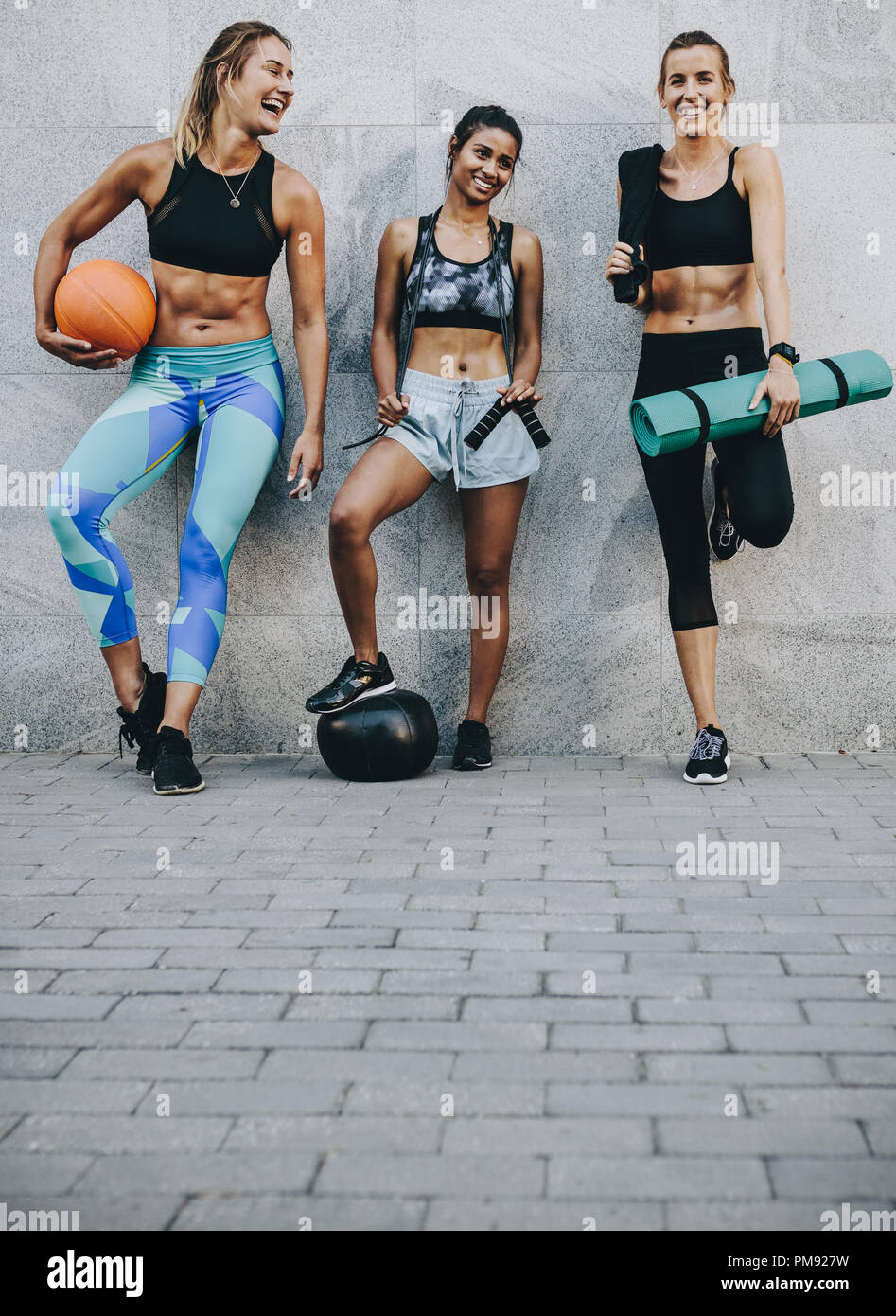 Sorridenti donne fitness in piedi in strada contro una parete tenendo la pallacanestro corda e il tappetino dopo allenamento. Foto Stock
