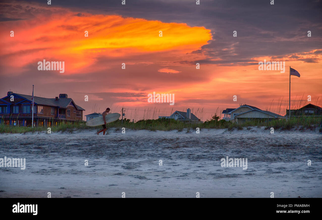 Giugno 18, 2018 Wrightsville Beach, NC: un surfista passeggiate lungo la sabbia sulla Wrightsville Beach come il sole tramonta in background. Foto Stock