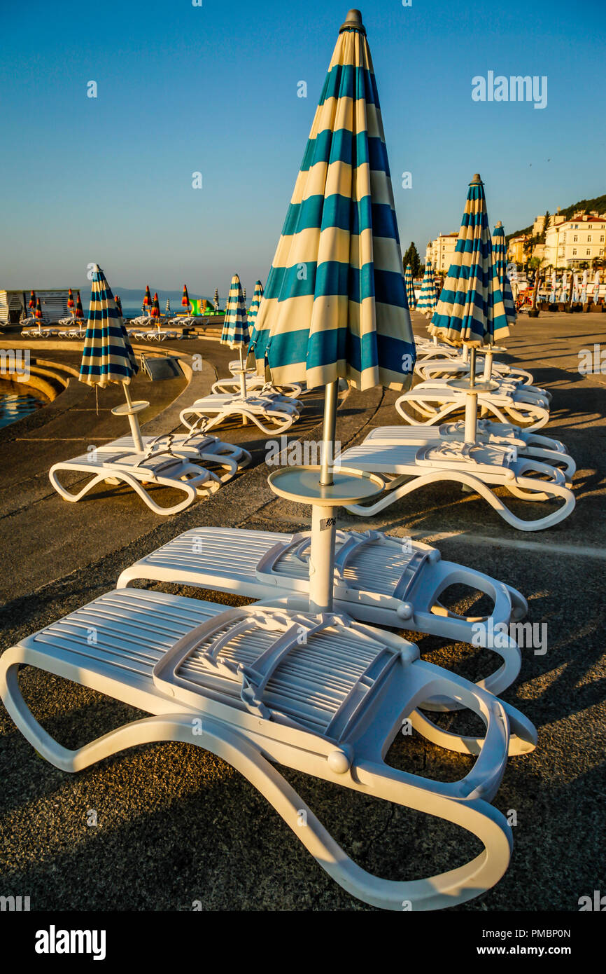 Svuotare sdraio in spiaggia con grandi piegata verso il basso ombrelloni linea il lungomare al tramonto in Opatija sul mare Adriatico in Croazia Foto Stock