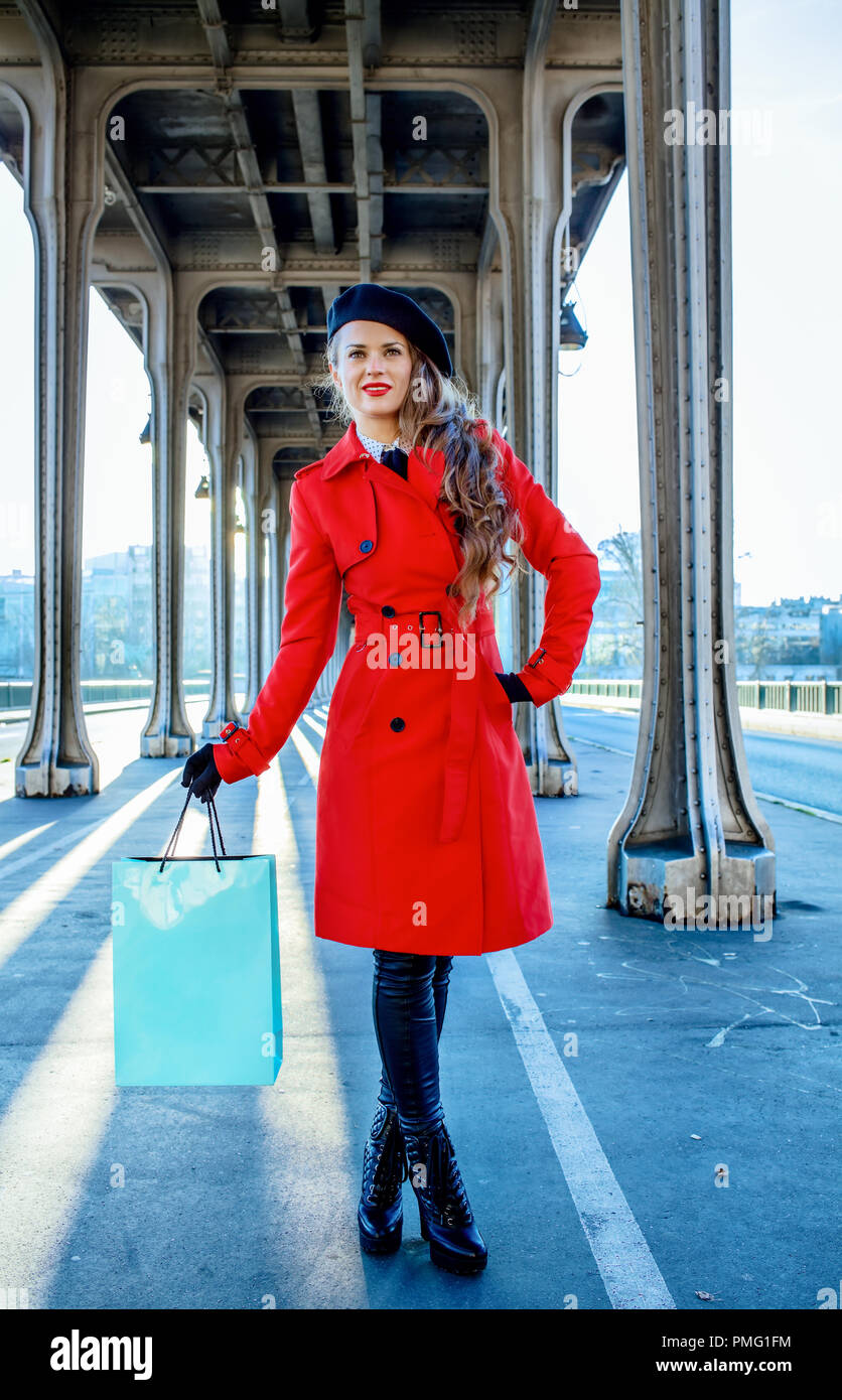 Luminose a Parigi. A piena lunghezza Ritratto di giovane viaggiatore woman in red trench coat sul Pont de Bir-Hakeim bridge in Paris holding shopping bag Foto Stock
