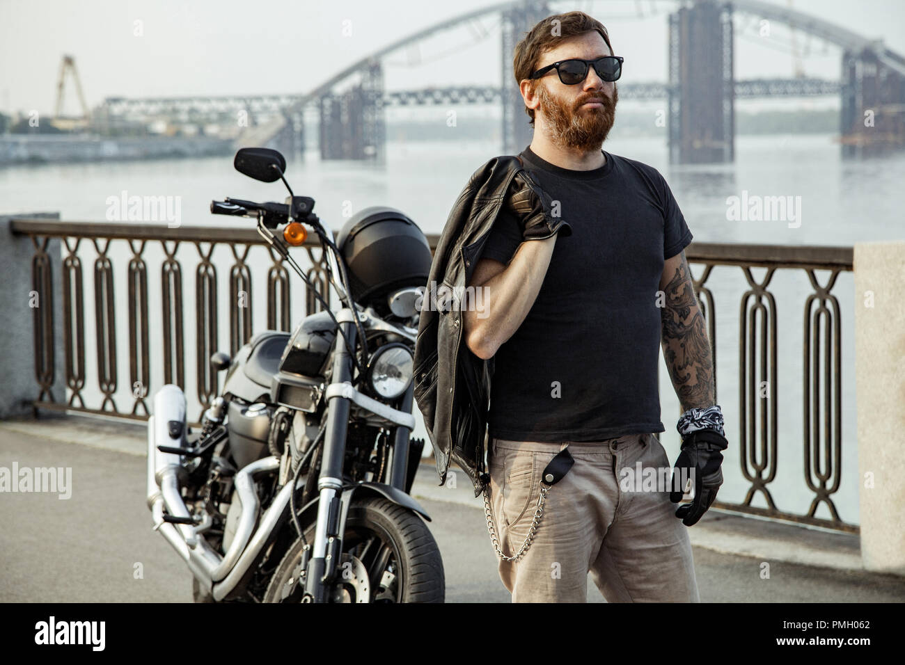 Redhead barbuto biker holding giacca di pelle sulla spalla e piedi vicino la moto sul ponte Foto Stock