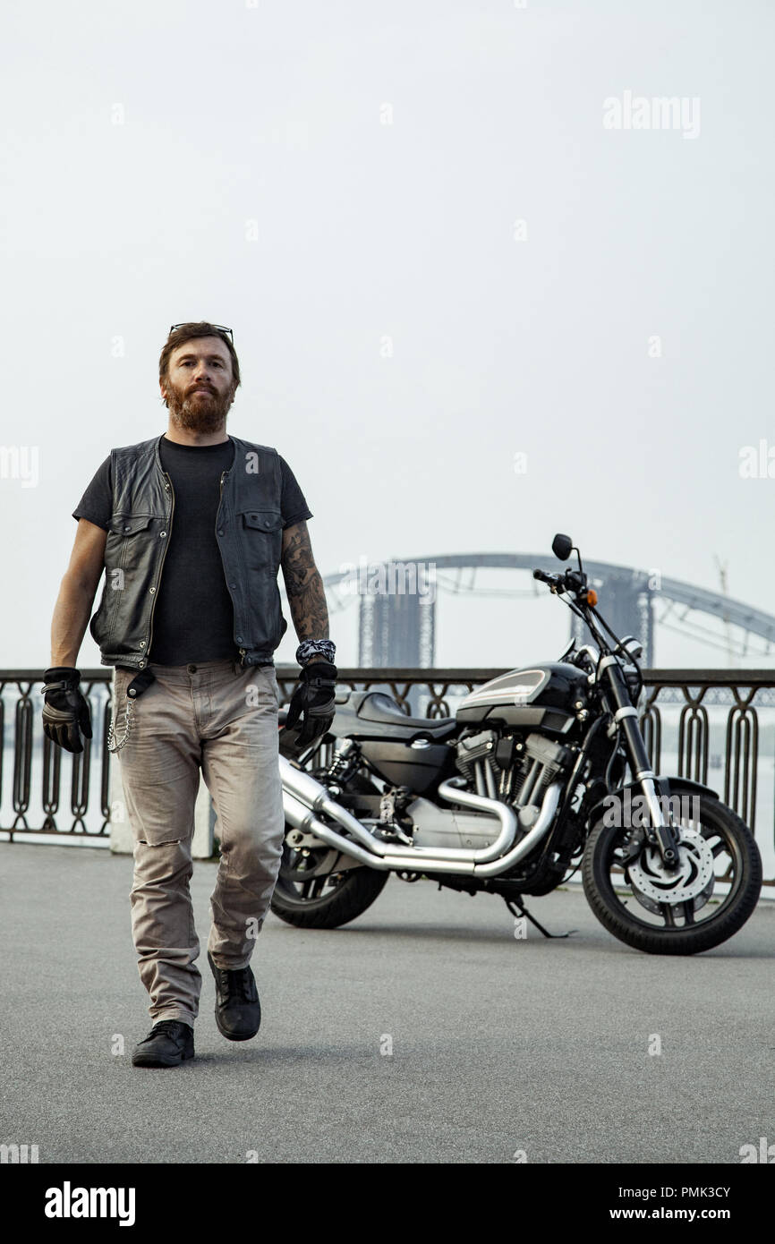 Redhead biker con la barba in giacca di pelle in piedi camminando sul ponte in prossimità di moto Foto Stock