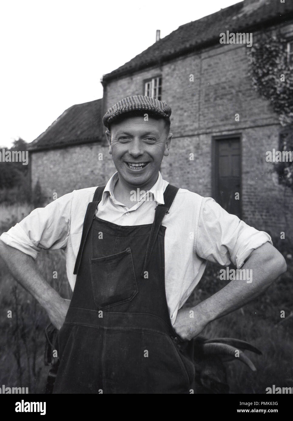 Degli anni Cinquanta, storico ritratto di un maschio di manutenzione immobiliare uomo o tuttofare in piedi al di fuori di una proprietà farm, indossando i suoi abiti da lavoro, maglietta a collare, dunagarees e tappo piatto, Inghilterra, Regno Unito. Foto Stock