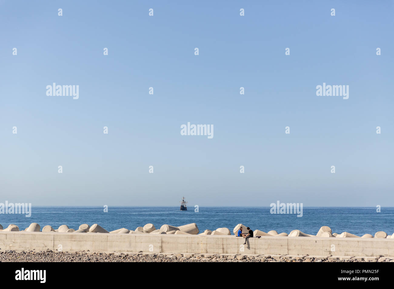 Coppia eterosessuale appendere fuori, seduti insieme sulla parete del mare, con molto il mare blu e una nave a vela in distanza Foto Stock