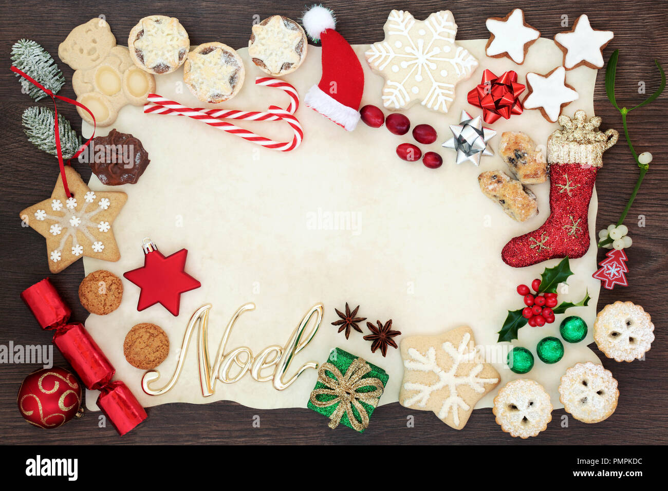 Natale e Noel del bordo dello sfondo compresi addobbi per l'albero, biscotti, torte, frutta, cioccolatini e flora invernale su carta pergamena sul rovere rustico. Foto Stock