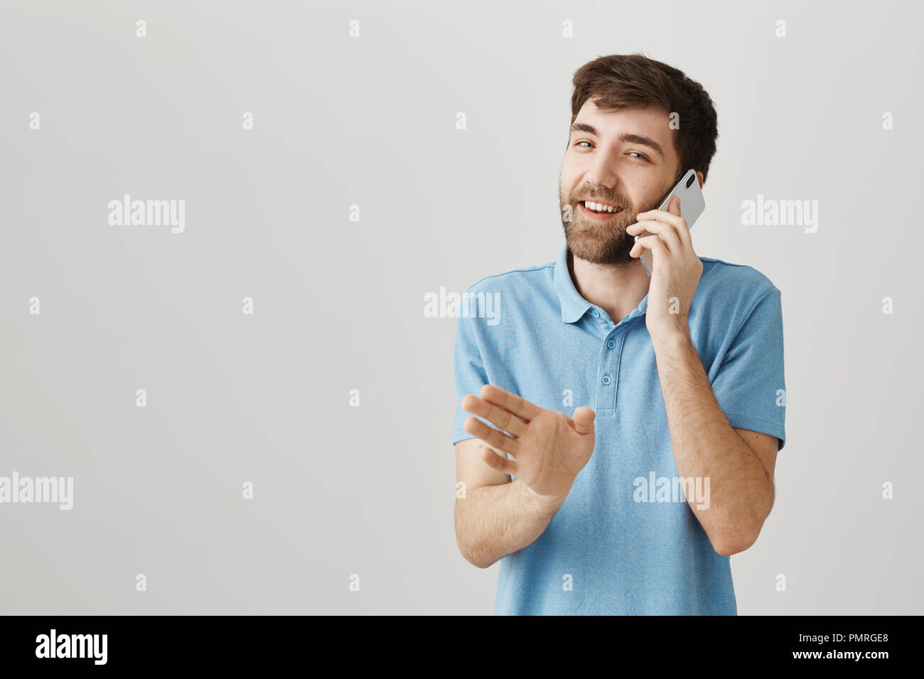 Appendere su ho bisogno di ordinare da bere. Ritratto di occupato e felice ragazzo caucasico parlando sullo smartphone, sorridere mentre sventolando in telecamera come se dicendo di attendere un secondo, in piedi su sfondo grigio Foto Stock