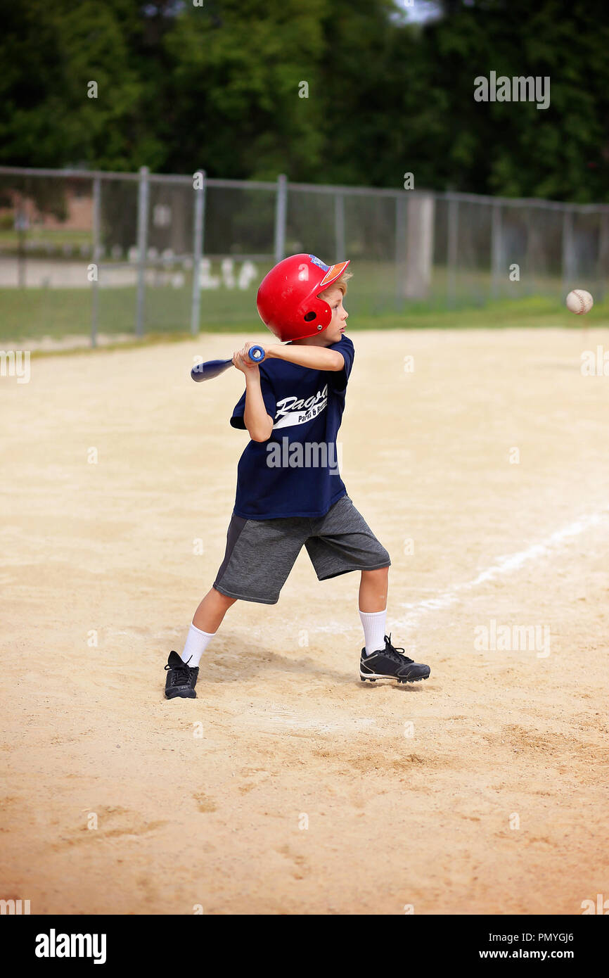 Un bambino di 8 anni giovane ragazzo indossa un casco ed oscillante di una mazza da baseball alla sfera in una gioventù ragball gioco in una piccola città. Foto Stock