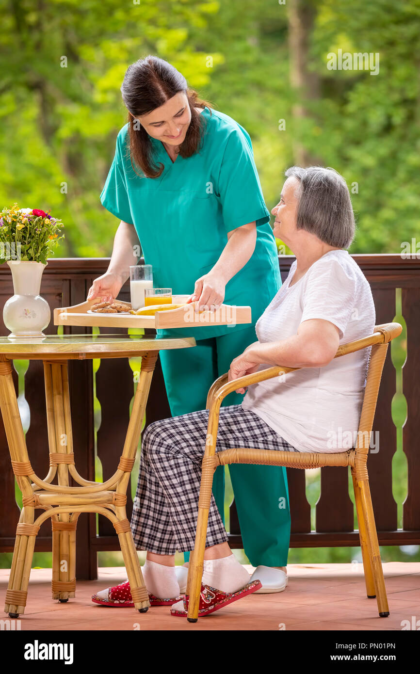 Felice l'infermiera o il caregiver che serve colazione sana per donna anziana sulla terrazza presso la casa di cura. Foto Stock