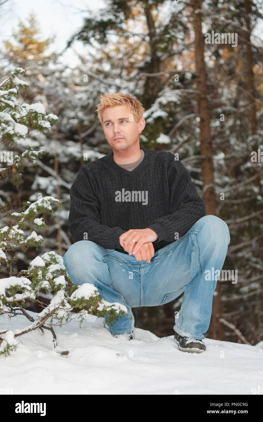 Uomo seduto accovacciato in snow all aperto in inverno lo sguardo lontano Foto Stock
