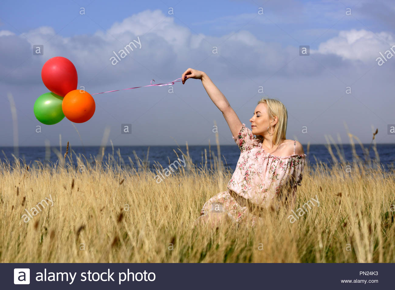Donna bionda in abiti estivi seduto in erba lunga giocando con palloncini Foto Stock