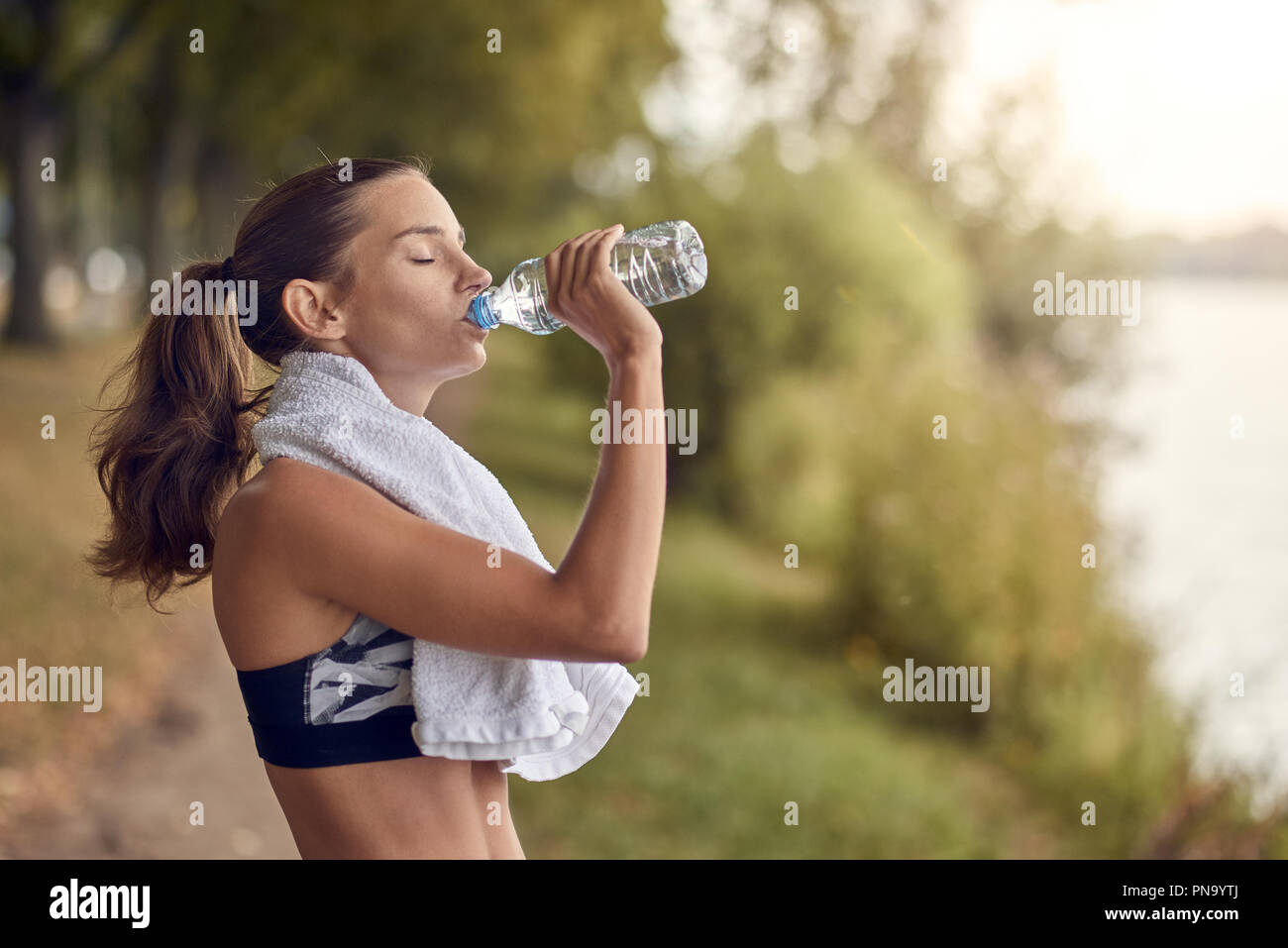 Montare sportivo da donna di bere acqua da una bottiglia di plastica come lei si interrompe durante il suo allenamento jogging su un viale alberato sentiero con un asciugamano intorno al collo Foto Stock