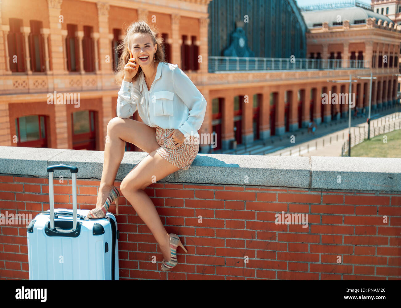 Ritratto di sorridere eleganti donna turistica con trolley borsa nella parte anteriore della Puerta de Atocha mentre è seduto sul parapetto Foto Stock