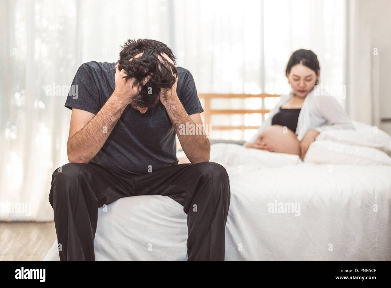 Preoccupato stress uomo seduto sul letto con la mano sulla fronte in camera da letto in gravi umore emozione con moglie incinta Donna sfondo. Depressivi maggiori Disor Foto Stock