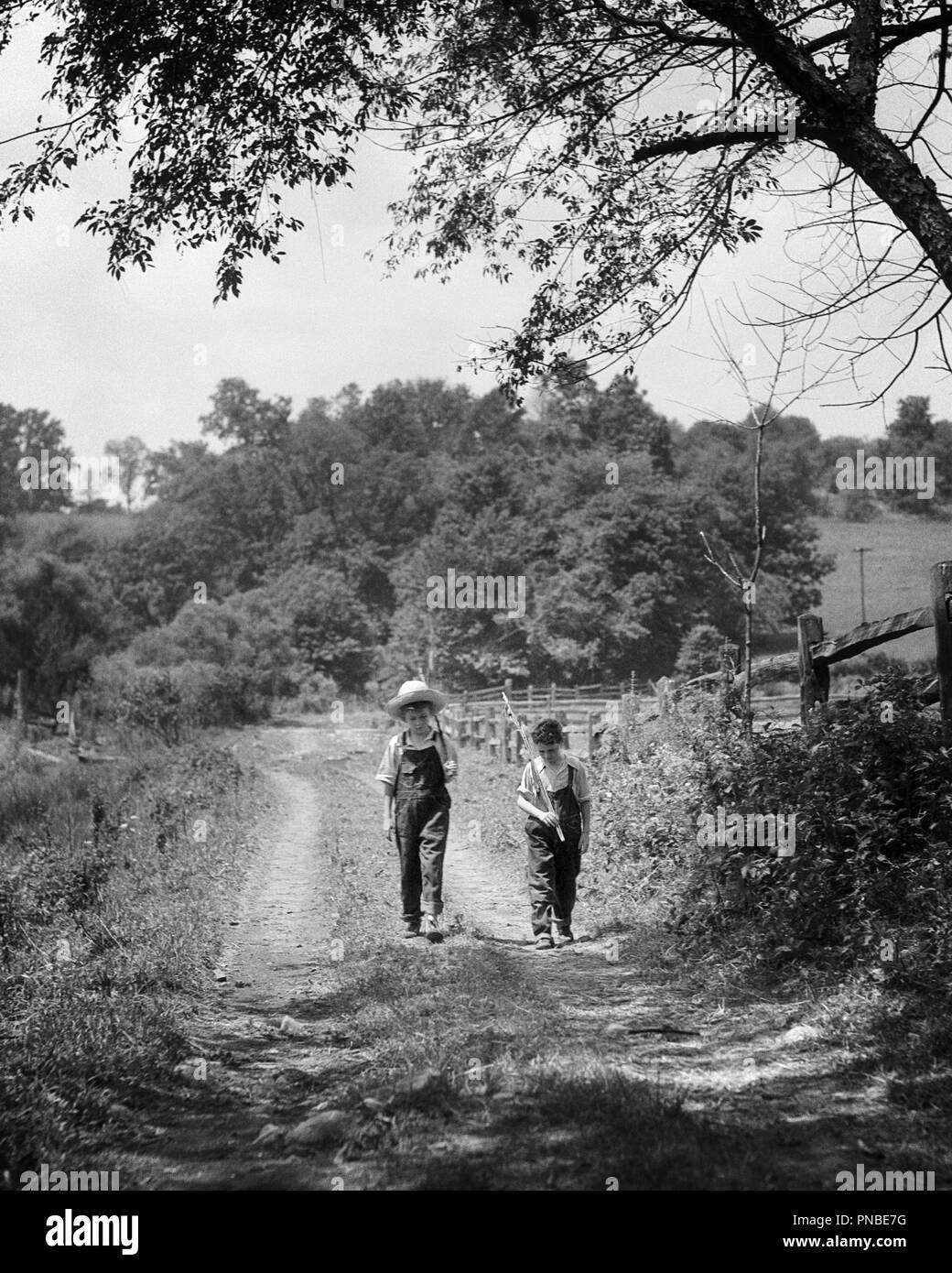 Negli anni Trenta due ragazzi camminare in Vicolo del paese trasportano STICK CANNE DA PESCA SULLA LORO SPALLA indossando tute da BIB - un2492 HAR001 HARS ANGLING BIB TUTE pre-teen pre-teen BOY aste convivere in bianco e nero di etnia caucasica HAR001 in vecchio stile Foto Stock