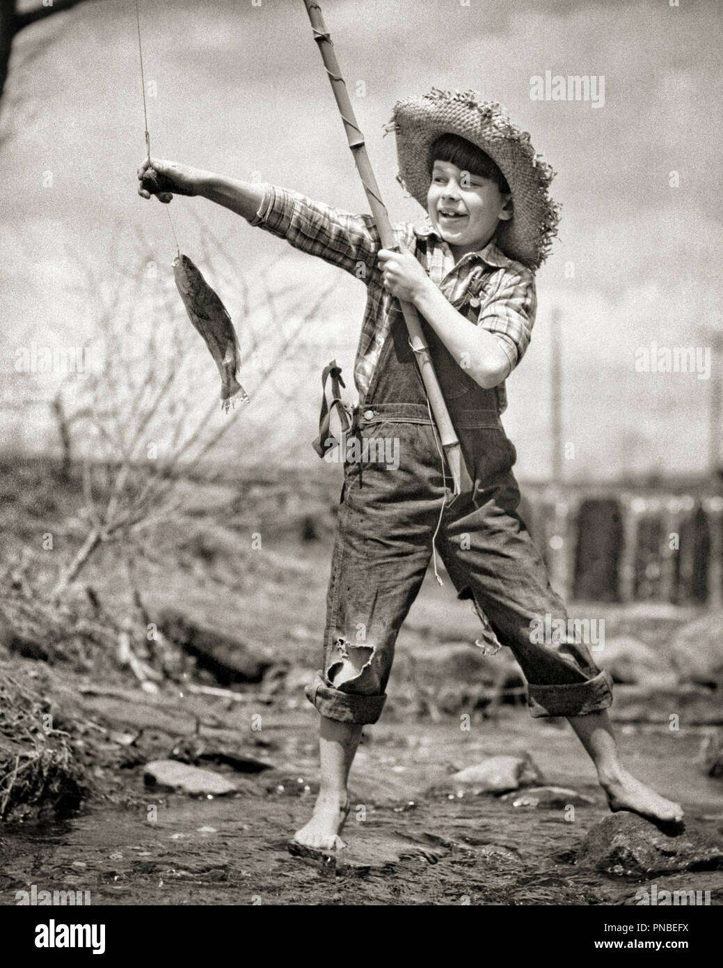 Negli anni quaranta anni cinquanta piedi nudi ragazzo in tuta nel flusso palo di bambù tenendo il pesce indossando cappello di paglia fori nelle ginocchia slingshot in tasca - una3216 DEB001 HARS ORGOGLIO NEL FINO A PIEDI NUDI SLINGSHOT FORI Huck Finn tute da BIB DEB001 Tom Sawyer Huckleberry Finn novellame BIANCO E NERO etnia caucasica in vecchio stile Foto Stock