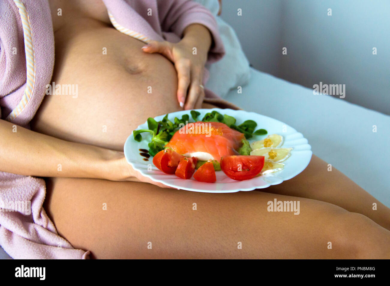 Piuttosto giovane donna incinta dalla pancia posa sul letto e tenendo una piastra con un sano cibo nutriente, gli acidi grassi omega 3 pasti deliziosi ingredienti Foto Stock