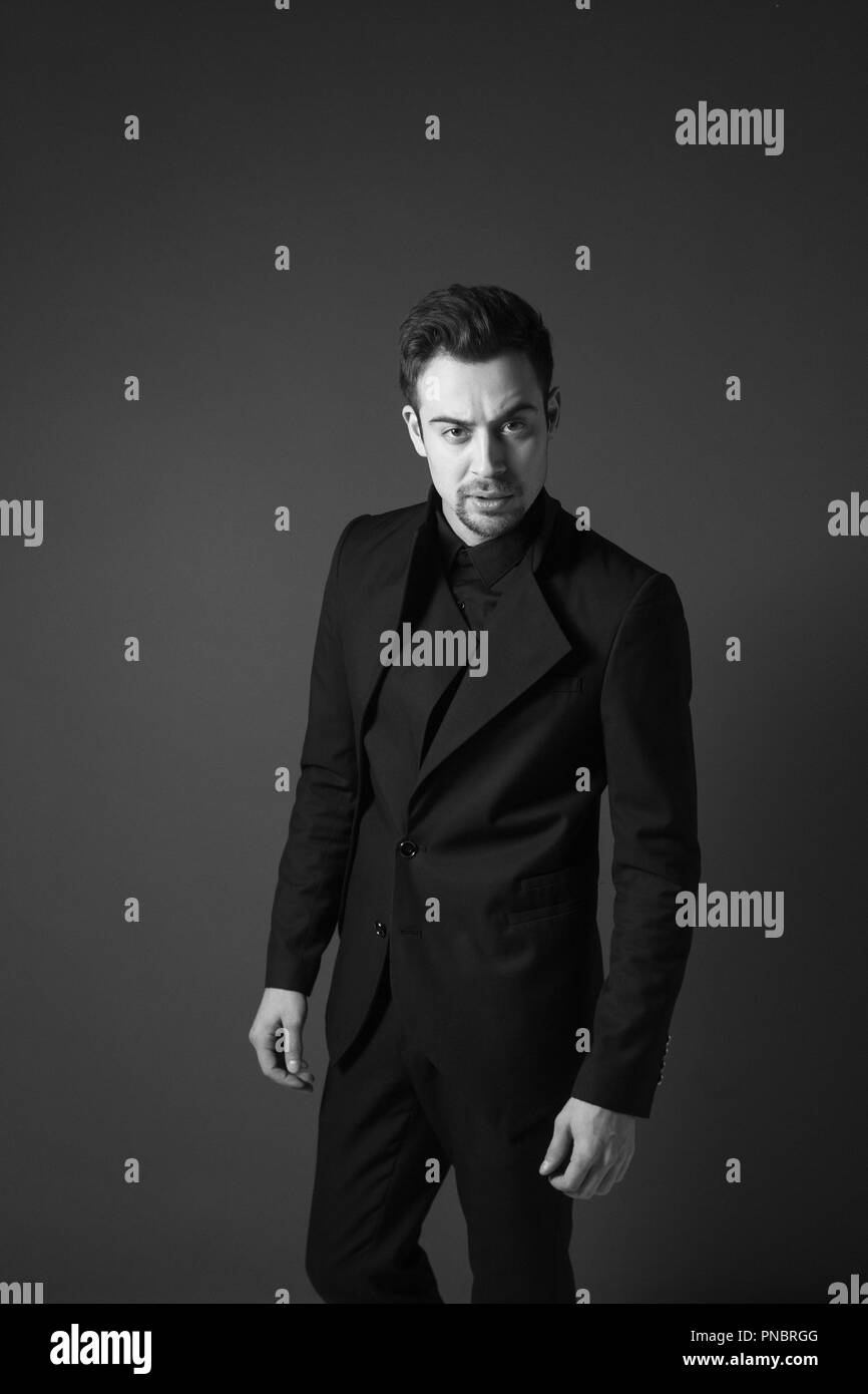 Giovane uomo bello in un vestito nero, in piedi e seriamente guardando la telecamera, contro la pianura di sfondo per studio Foto Stock