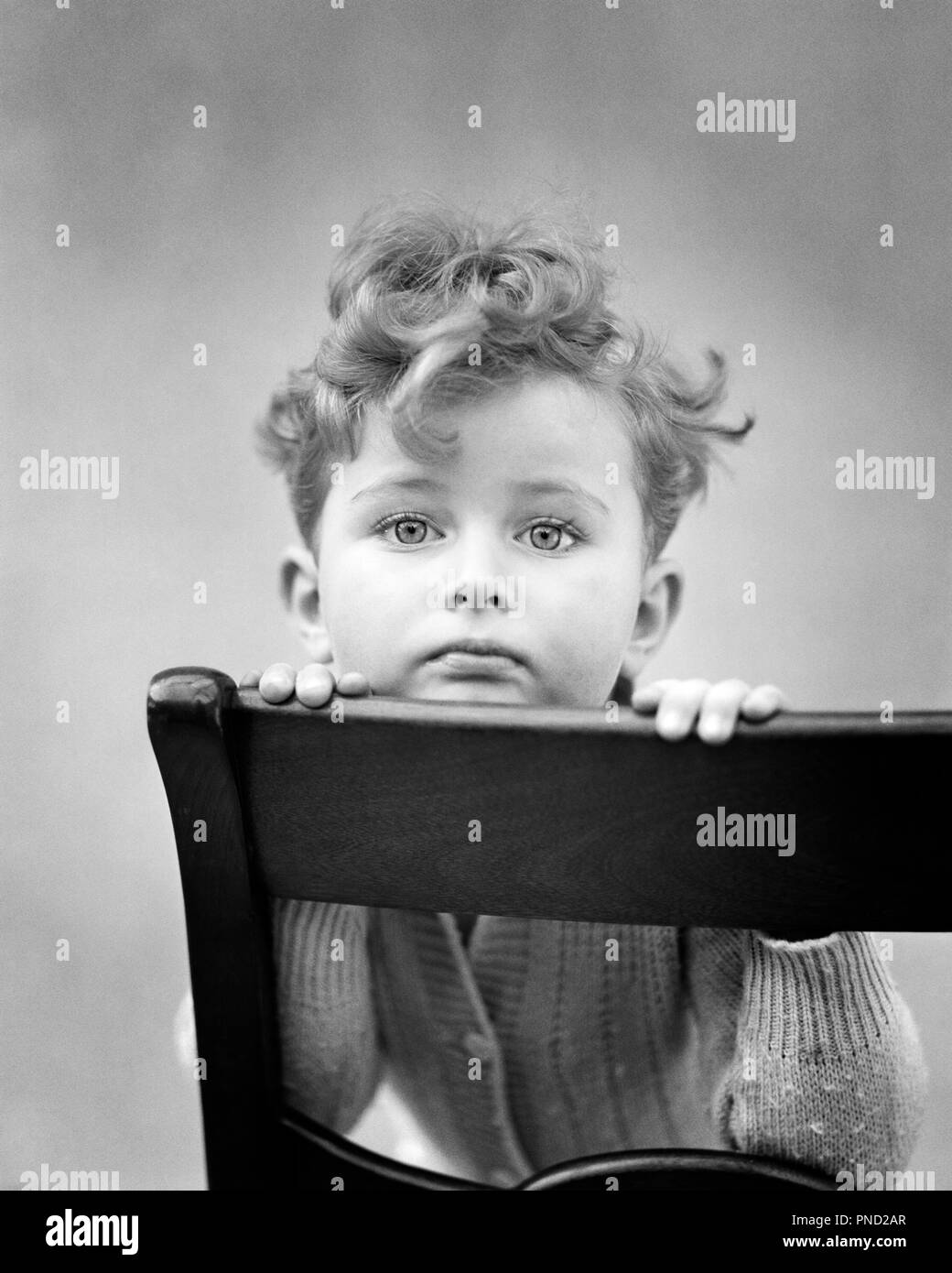 1940s diffidente nei capelli ricci BOY mento di appoggio sulla parte posteriore della sedia guardando telecamera - J9147 HAR001 HARS tristezza ampio contatto visivo ansia fissando la testa e spalle ansiosi RICCI DI fisso sulla sensazione di apprensione COLLEGAMENTO GLUM concettuale ritirati timidezza vacante diffidenti emozione emotiva crescita emozioni novellame misero in bianco e nero di etnia caucasica HAR001 in vecchio stile Foto Stock