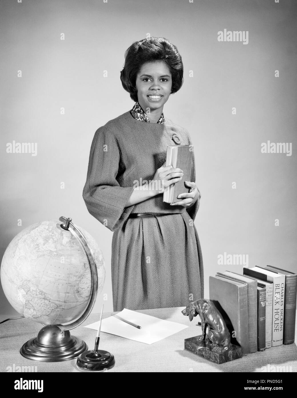 Anni sessanta sorridente americano africano donna insegnante presso la sua scrivania con i libri di testo guardando la fotocamera - n2061 HAR001 HARS B&W I sogni di felicità occupazione allegro avventura afro-americani afro-americano carriere esperto etnia nero orgoglio occupazioni sorrisi libri di testo gioiosa dedizione elegante esperienza giovane donna adulta IN BIANCO E NERO DEDICATA HAR001 in vecchio stile afro-americani Foto Stock