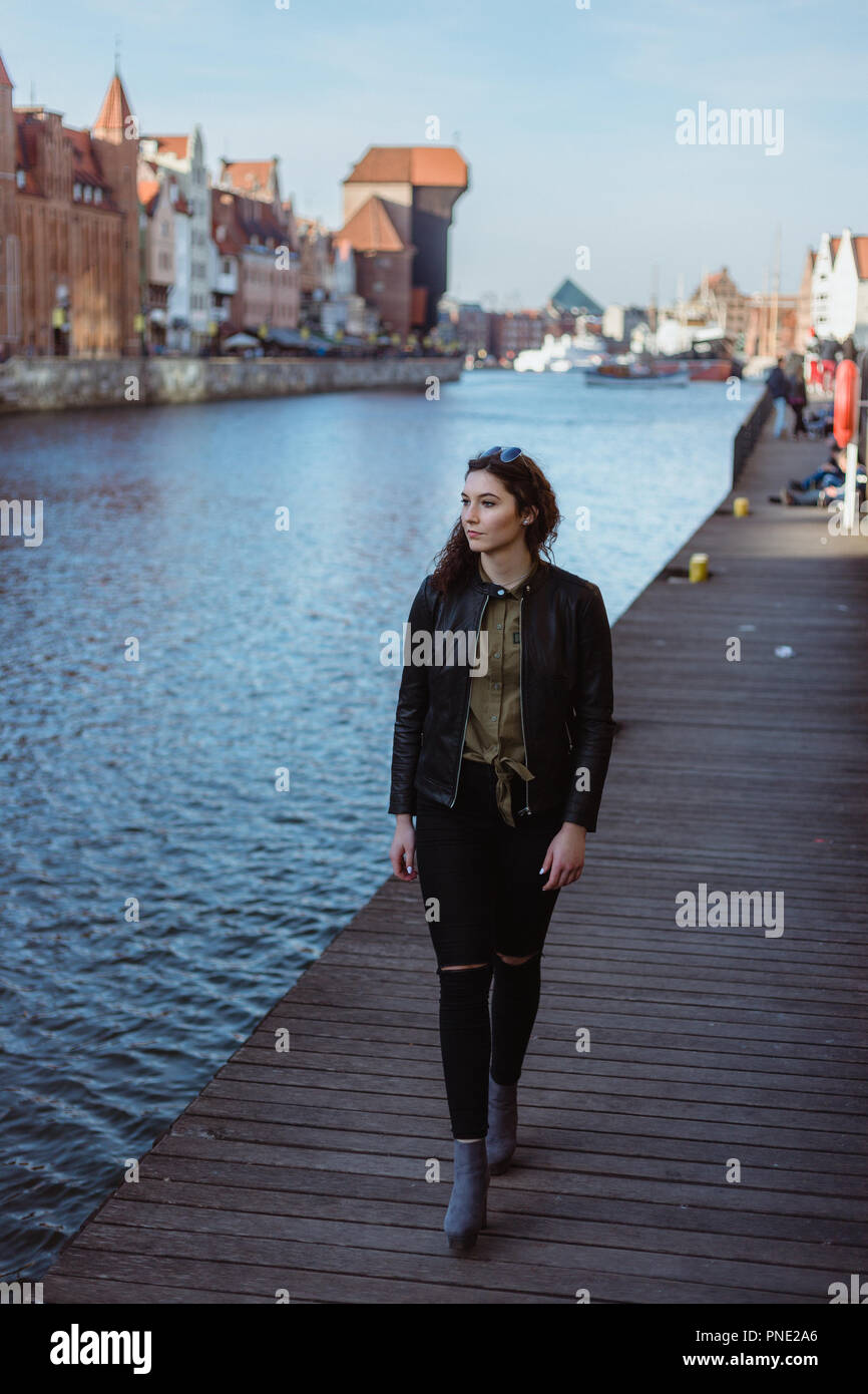 Giovane donna che indossa una giacca di pelle è a piedi dal molo sul fiume in Gdansk, Polonia, sorridente e guardando verso il basso. Foto Stock