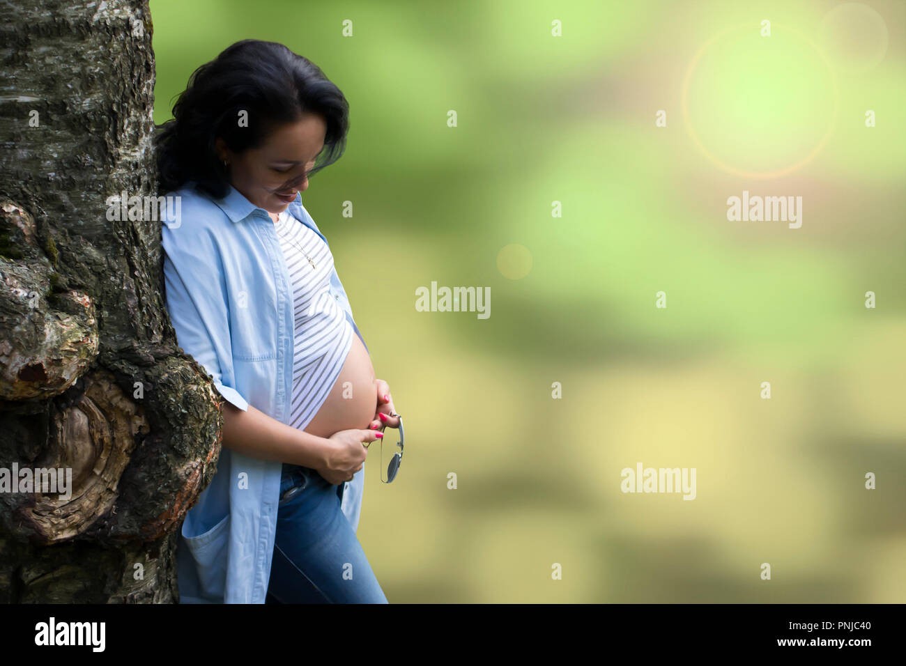 Giovane bella capelli lunghi brunette appoggiato su di un albero di bizzarro e guardando la sua gravidanza ventre contro sfocato sfondo bokeh di fondo Foto Stock