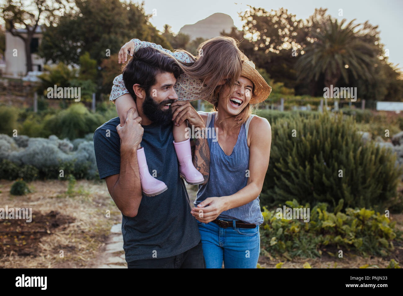 Barba uomo che porta bambina sulla spalla con la donna che ride presso l'azienda. Bella famiglia di tre divertirsi presso la loro fattoria. Foto Stock