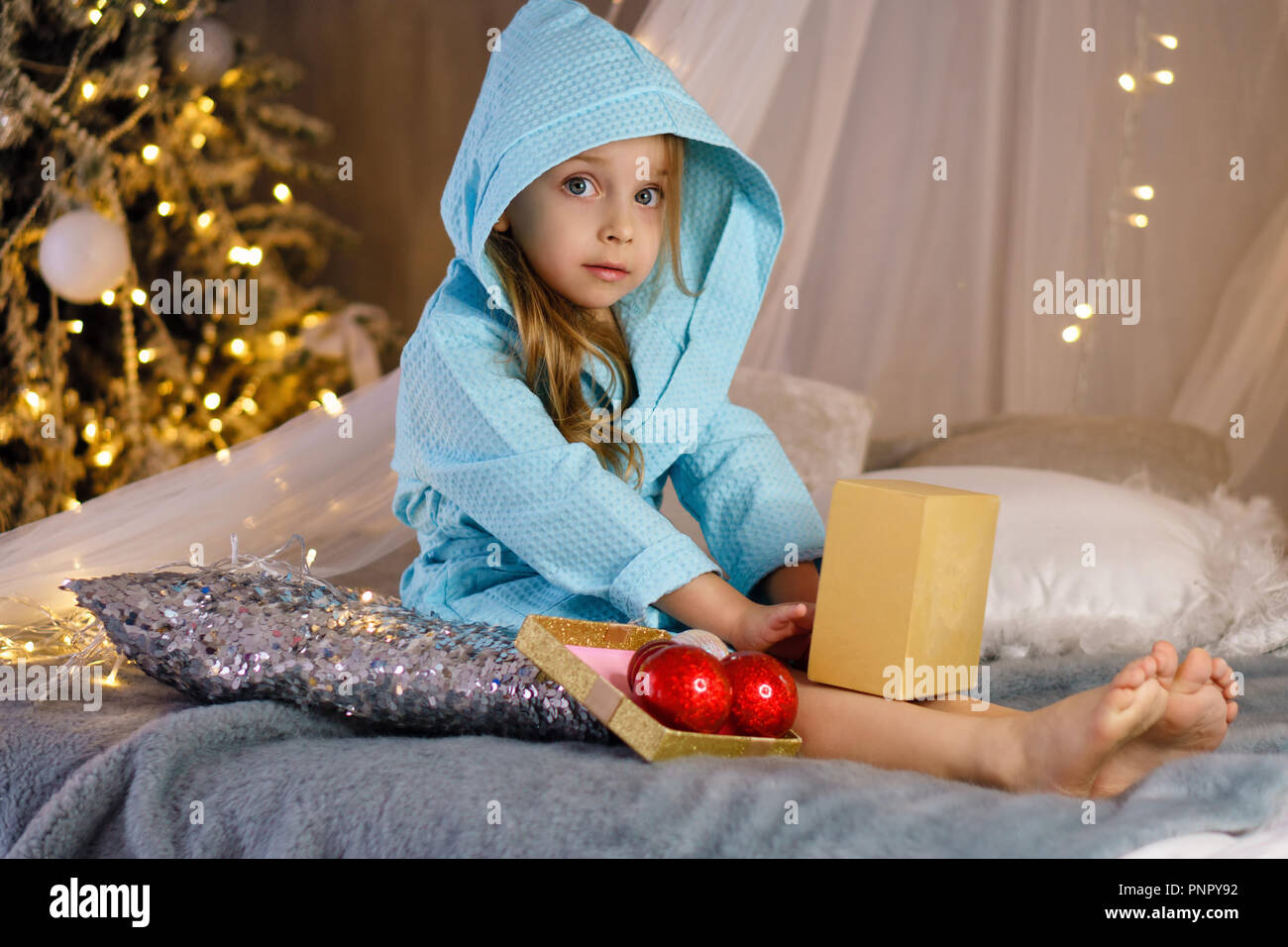 Bambina in accappatoio è seduta sul letto. Albero di natale con ornamenti in background. Infanzia felice. Foto Stock