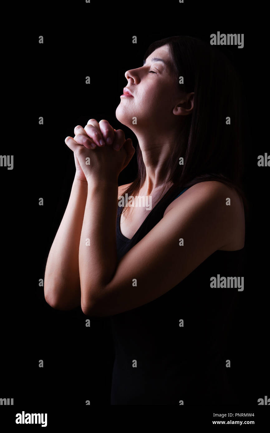 Fedeli donna orante, mani giunte nel culto a Dio con la testa in alto e gli occhi chiusi in fervore religioso, su uno sfondo nero. Concetto di religione, Foto Stock