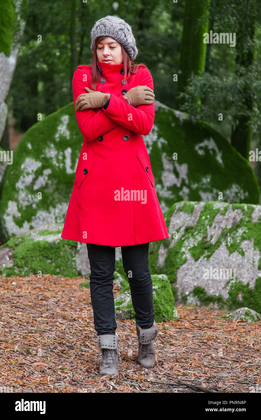 Giovane donna perso su una foresta i brividi di freddo e abbracciando o azienda stessa, indossando un rosso lungo cappotto o soprabito, un beanie e guanti durante la fal Foto Stock