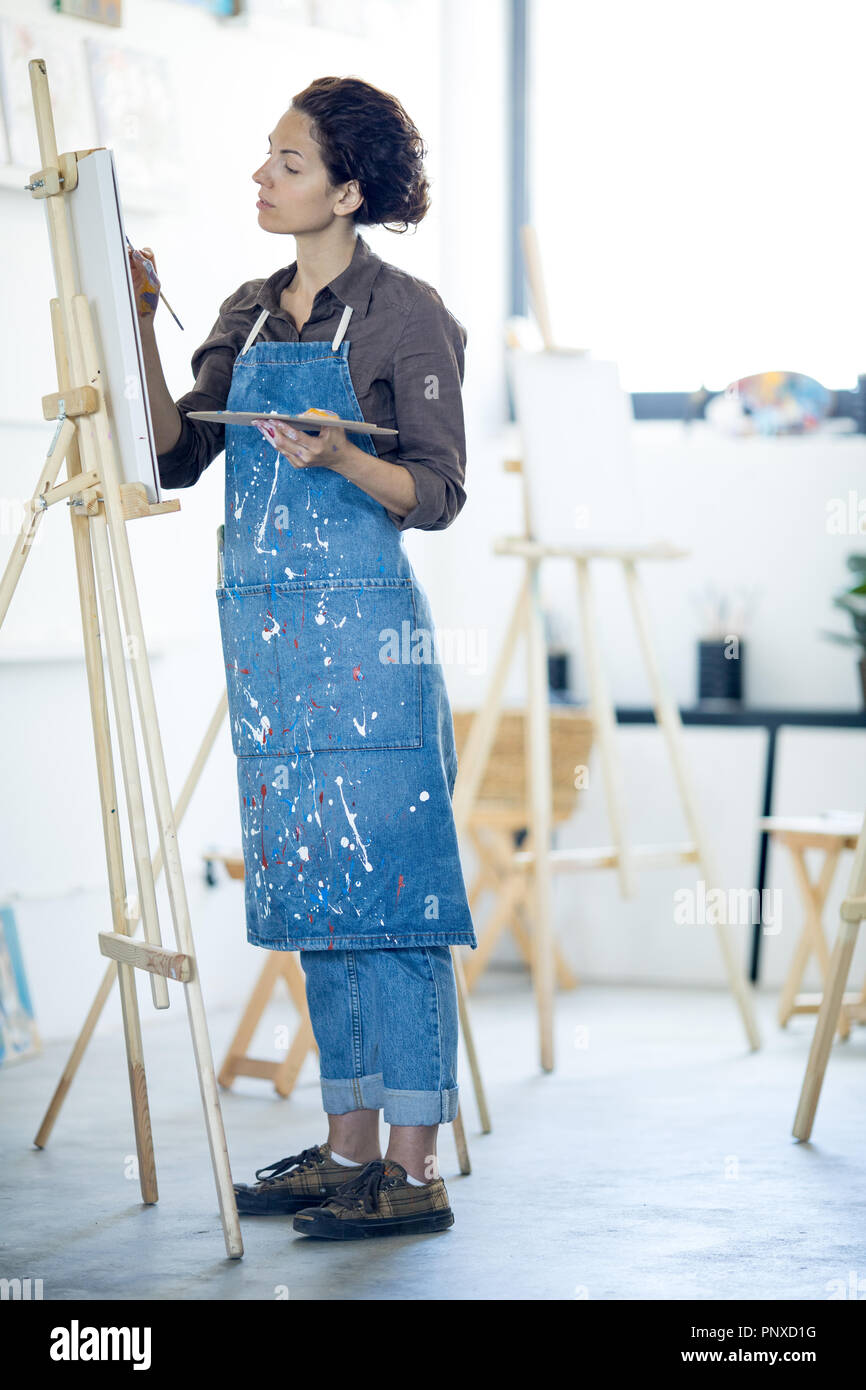 Giovani donne in abbigliamento da lavoro in piedi dal cavalletto mentre la pittura in opere di arte moderna studio Foto Stock