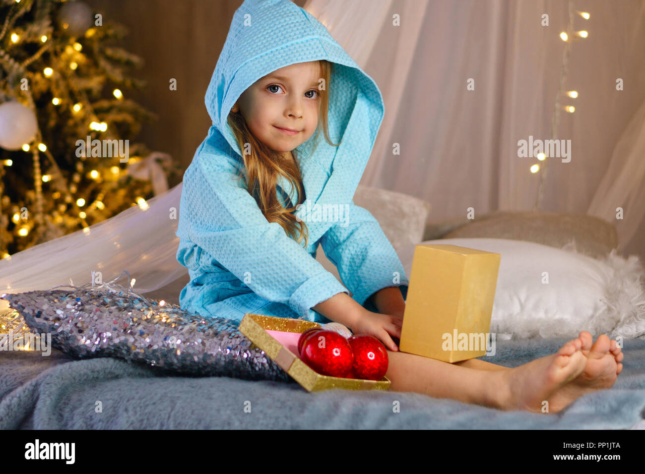 Bambina in accappatoio si siede sul letto e sorrisi. Albero di natale con ornamenti in background. Infanzia felice. Foto Stock