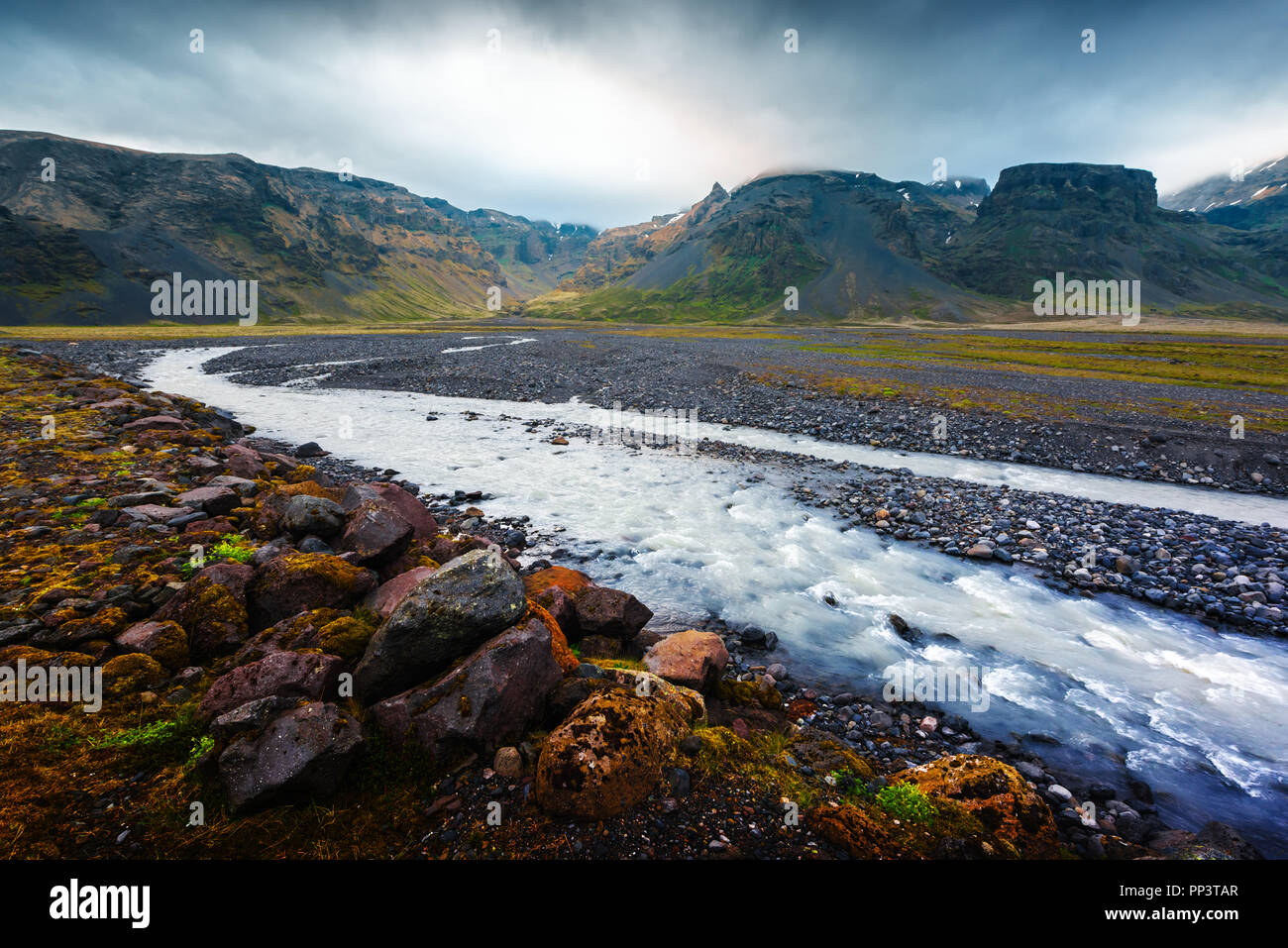 Islanda tipico paesaggio con fiume e le montagne. Cielo drammatico e pietre rosse su un primo piano Foto Stock