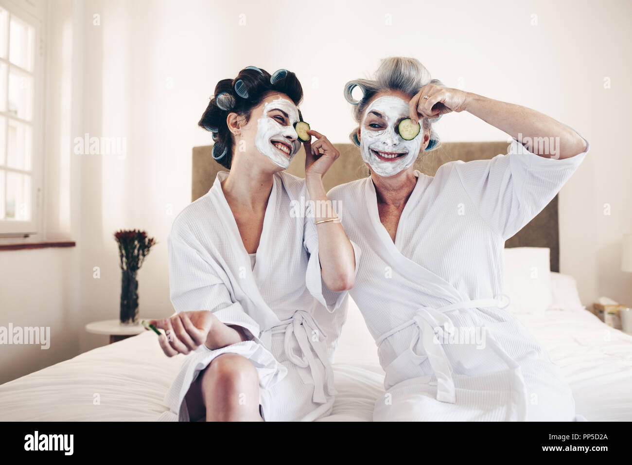 Due donne sorridenti tenendo una fetta di cetriolo ai loro occhi con una faccia pack sul viso. Madre e figlia in accappatoi seduta sul letto con rulli pinn Foto Stock