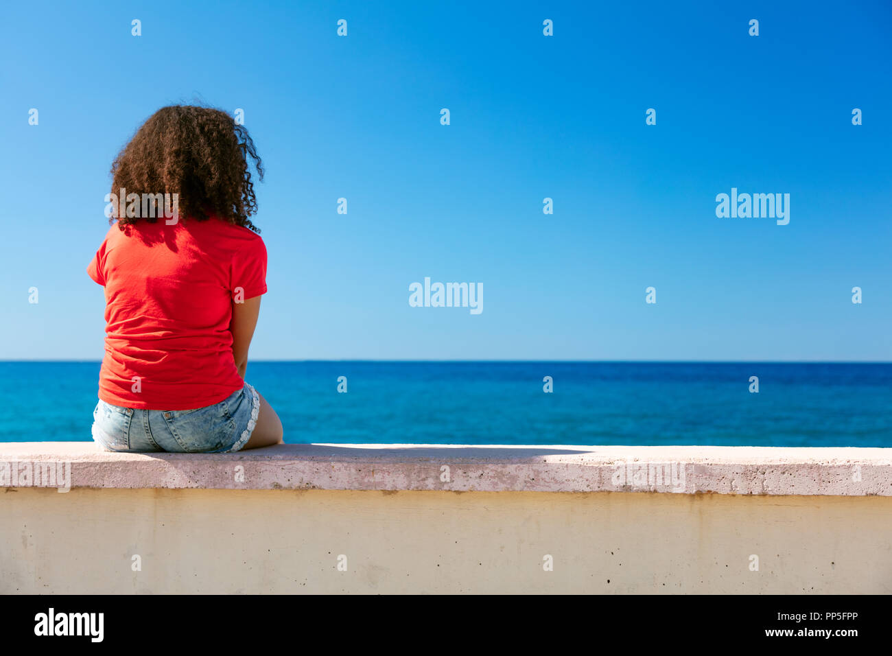 Razza mista americano africano ragazza adolescente femmine giovane donna che indossa una t-shirt rossa e gli shorts in denim seduto su una parete che guarda a un mare blu Foto Stock