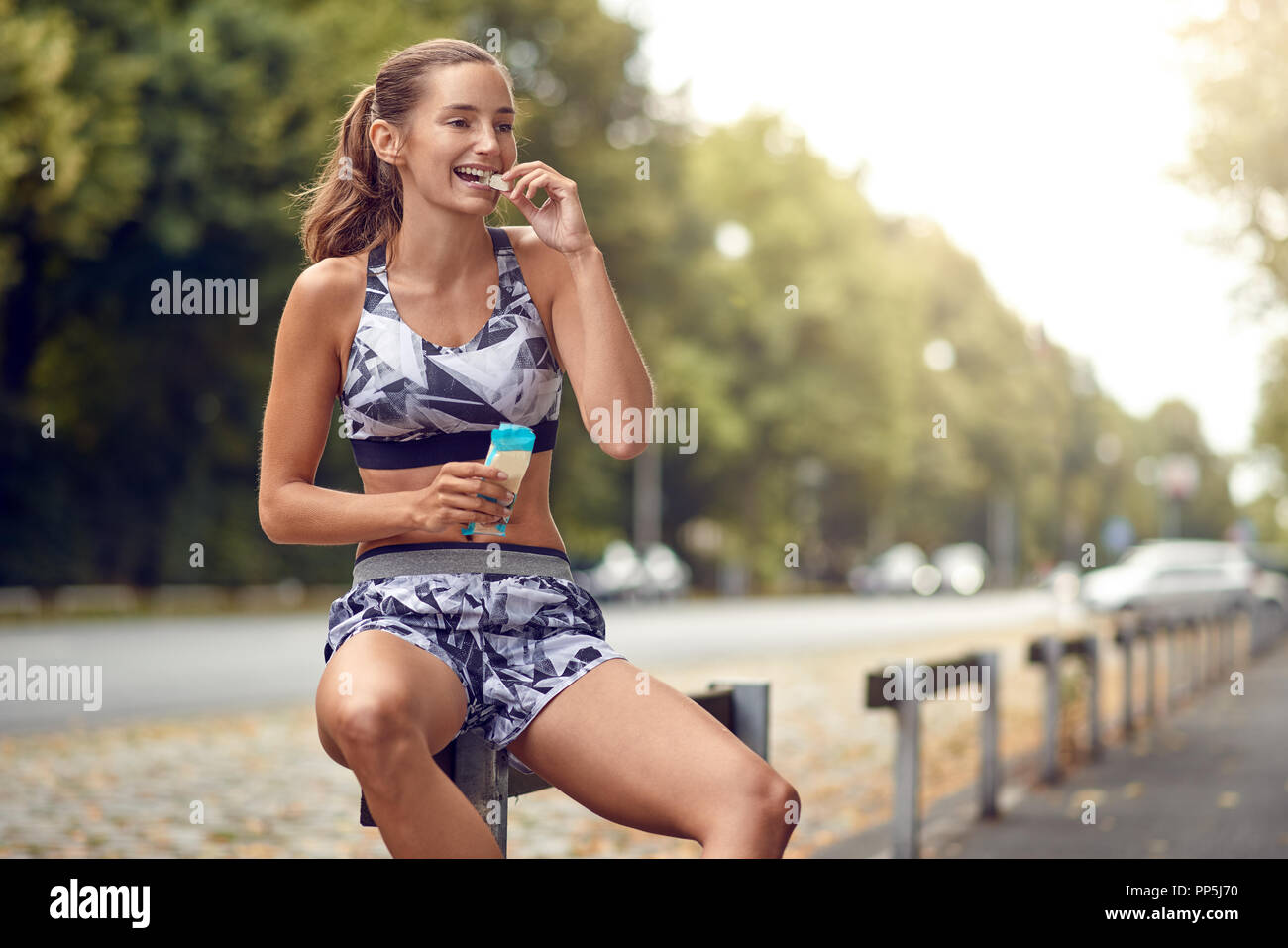 Attraente sportivo snello giovane donna seduta su una rampa stradale di mangiare un pezzo di una proteina bar come ella si prende una pausa dalla sua quotidiana jog Foto Stock