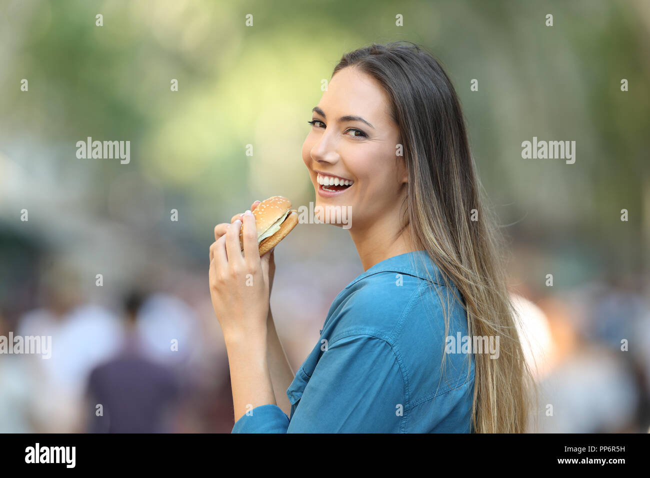 Donna felice di mangiare un hamburger guardando la telecamera in strada Foto Stock