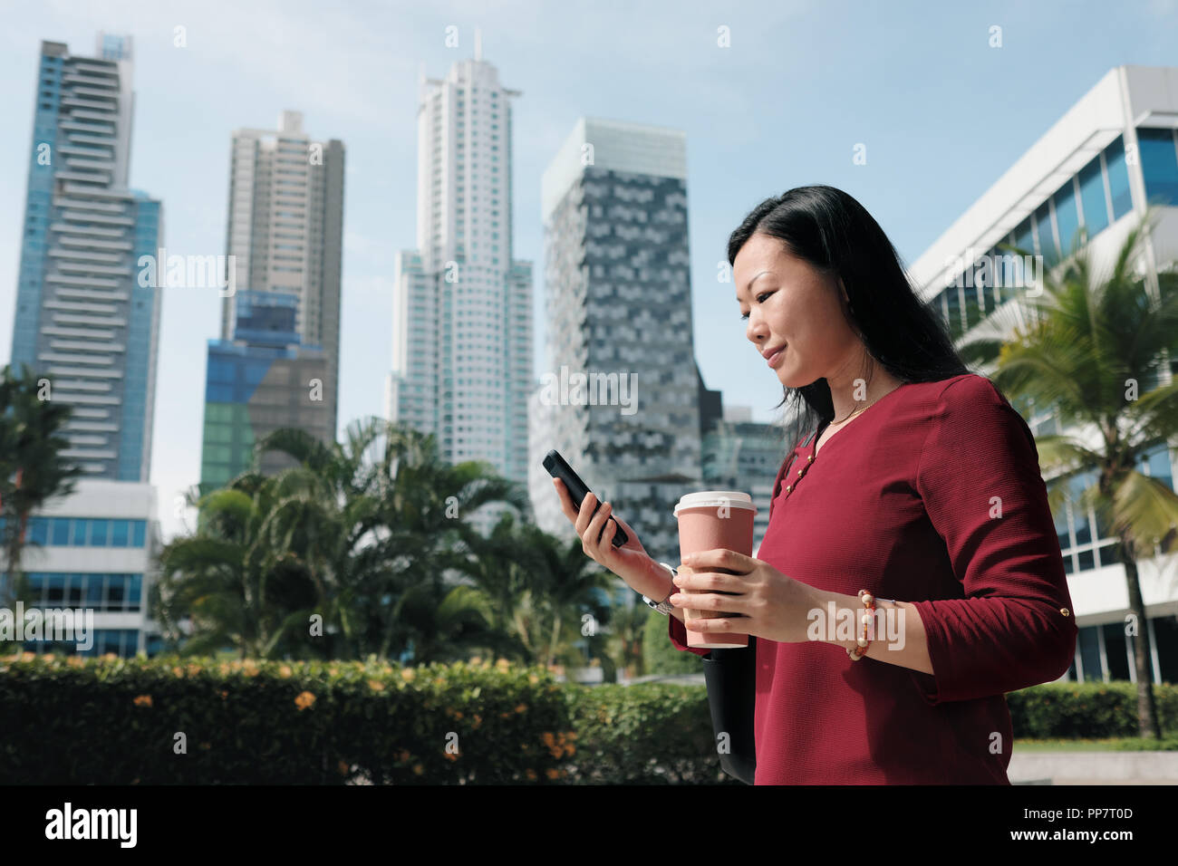 Felice femmina cinese manager messaging sul telefono cellulare, a bere caffè. Successo imprenditrice asiatica è raggiungibile a piedi in ufficio al mattino, occupato lady inviare Foto Stock