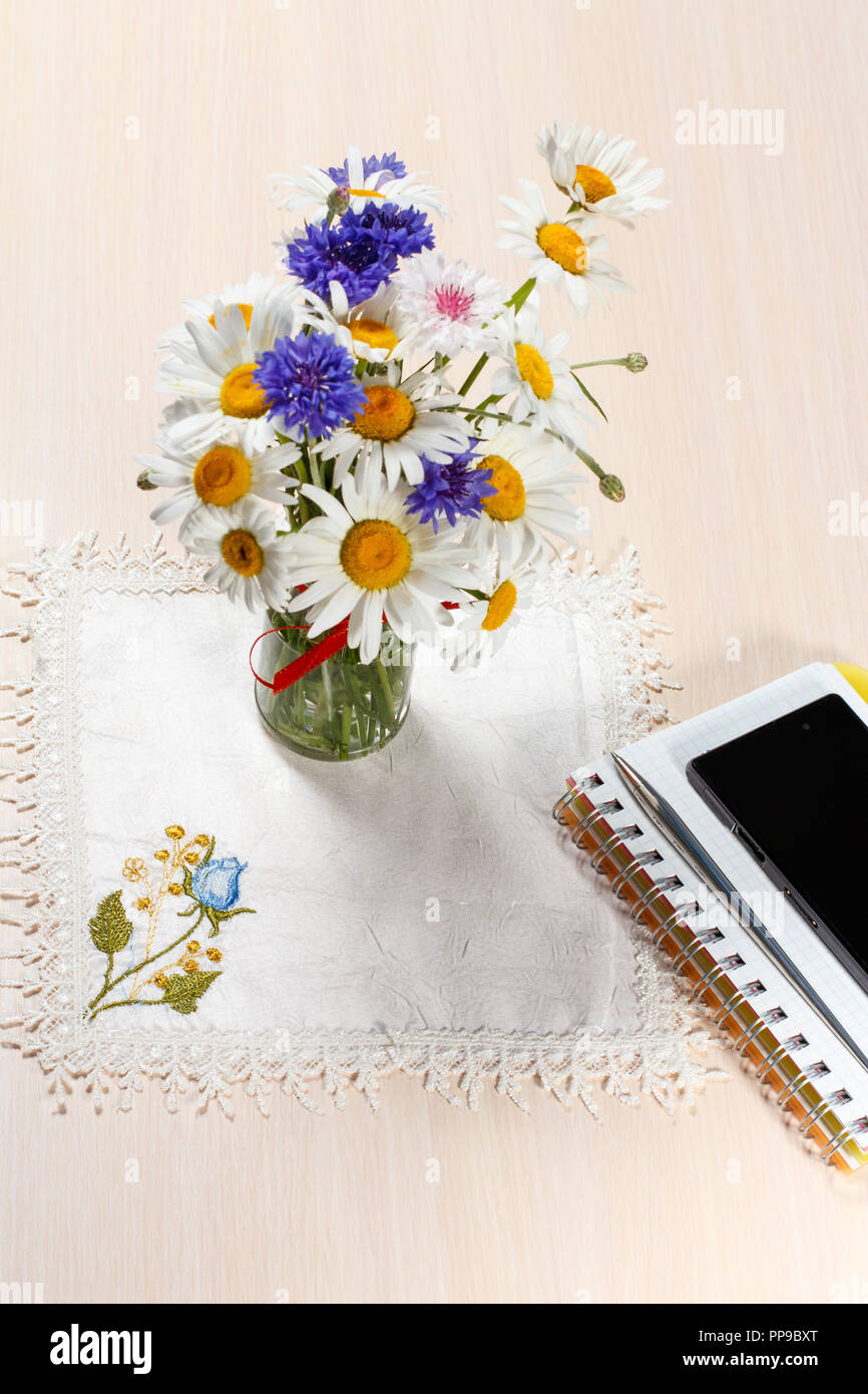 Daisy camomilla e cornflowers in vaso su un tavolo di legno con un tovagliolo, notebook, cellulare e una penna Foto Stock