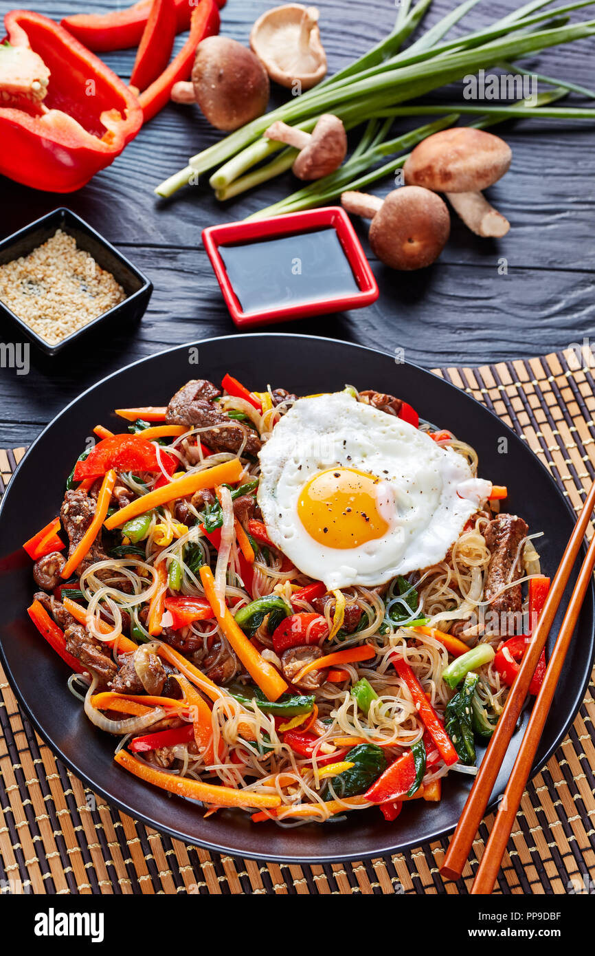 Il coreano mescolare noodles fritti e verdure con carne di manzo, funghi shiitake, guarnita con Sun su lato uovo, con semi di sesamo tostati, vestito con il mix o Foto Stock