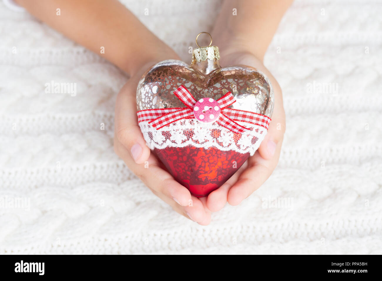 Bambino tiene un cuore di vetro, un giocattolo di Natale nelle mani sullo sfondo di un maglione caldo. Foto Stock
