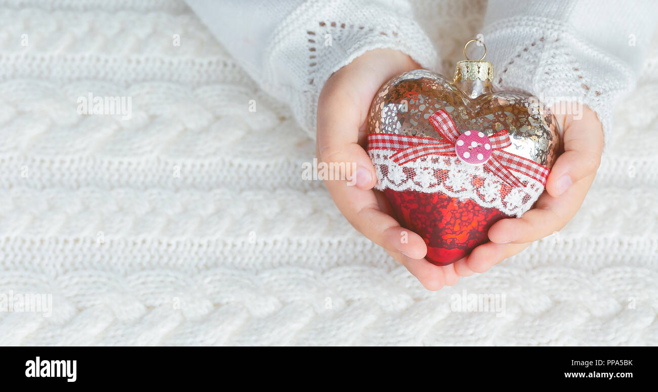 Bambino tiene un cuore di vetro, un giocattolo di Natale nelle mani sullo sfondo di un maglione caldo. Panorama Foto Stock